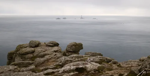 This is a landscape photograph taken in the early evening during the summer season, featuring the misty Longships lighthouse rocks as the main subject. The image captures the rugged cliffs of Cornwall in the foreground, overlooking the expansive and calm sea that stretches out towards the horizon. The Longships Lighthouse, located on the distant rocks, is visible through a veil of mist, emphasizing the atmospheric conditions typical of Cornwall’s coastline. The rocky terrain of the cliffs and the serene sea together highlight the characteristic natural beauty of this region.
